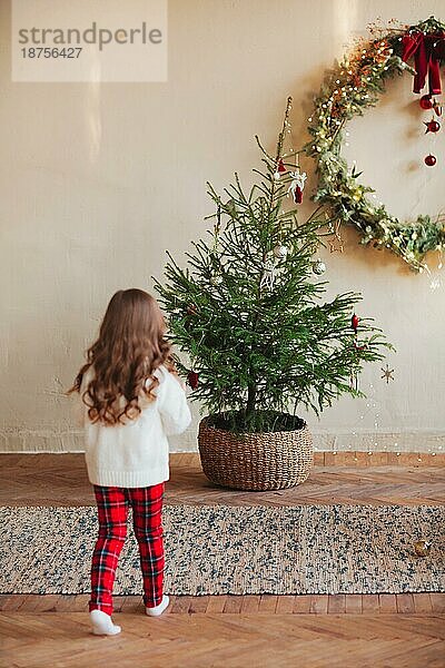 Nettes kleines Mädchen auf dem Teppich unter dem geschmückten Weihnachtsbaum während der Winterferien  trägt weißen geknöpften Pullover mit karierten Hosen