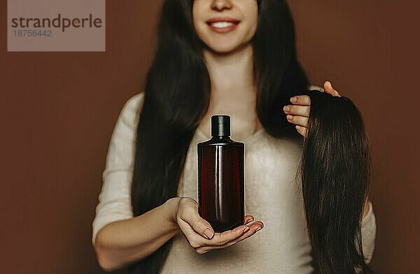 Junge lächelnde Frau mit langen gesunden braunen Haaren  die ein Shampoo mit organischen  natürlichen Inhaltsstoffen in der Hand hält. Glückliche Frau wirbt für Haarpflegeprodukte  während sie vor einem Studiohintergrund steht