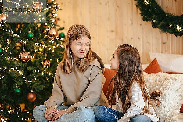 Gemütliche inländischen Porträt von zwei glücklichen lächelnden kleinen Mädchen Schwestern umarmt  während die Freizeit zusammen zu Hause zu verbringen  sitzen auf dem Bett in der Nähe geschmückten Weihnachtsbaum. Familie und Kinder Konzept