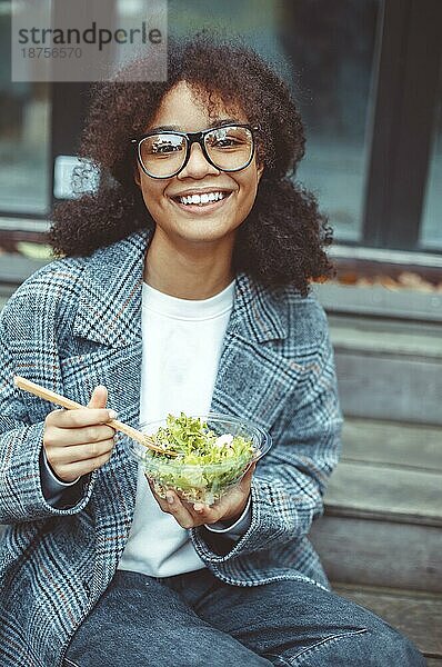 Glückliche afroamerikanische Frau Büroangestellte ißt Salat und lächelt in die Kamera  während sie auf einer Bank im Park im Freien sitzt  selektiver Fokus. Positives schwarzes Mädchen beim Mittagessen draußen während der Pause bei der Arbeit