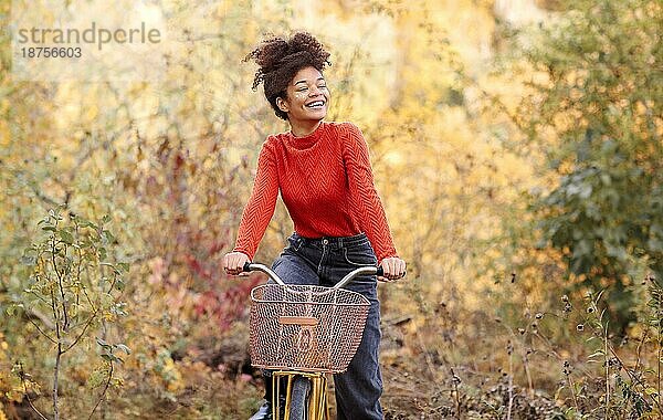 Junge glückliche lächelnde afroamerikanische Frau beim Fahrradfahren im Herbstwald  aktive positive gemischtrassige Frau in Freizeitkleidung  die in die Kamera lächelt  während sie mit dem Stadtrad in der herbstlichen Natur steht