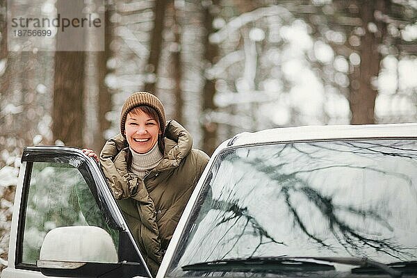 Fröhliche Frau lächelt und schaut in die Kamera  während sie ein modernes Fahrzeug auf einem unscharfen Hintergrund einer verschneiten Landschaft fährt