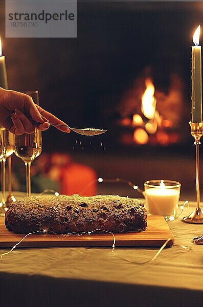 Weihnachtsessen. Weibliche Hand bestreut Puderzucker auf Weihnachten Kuchen Kuchen stehen auf rustikalen Holztisch mit Gläsern Champagner und brennenden Kerzen  vertikale Zusammensetzung. Selektiver Fokus. Stollen