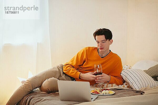 Entspannter junger Mann mit einem Glas Wein in der Hand  der auf einem bequemen Bett mit einem Teller Essen liegt und beim Essen Videos auf dem Laptop anschaut  während er sich zu Hause entspannt