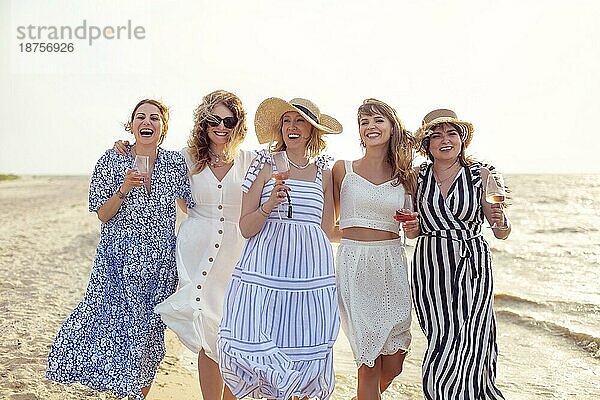 Gruppe von begeisterten Freundinnen mit Gläsern Wein lachend und umarmend beim Spaziergang am Strand am Sommertag zusammen