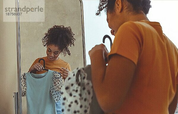 Junge lächelnde afroamerikanische Frau  die zu Hause vor dem Spiegel Kleidung anprobiert  glückliches schwarzes Mädchen  das ein blaues Sommerkleid auf einem Bügel hält und sich entscheidet  was sie für einen Spaziergang im Freien anziehen soll. Selektiver Fokus