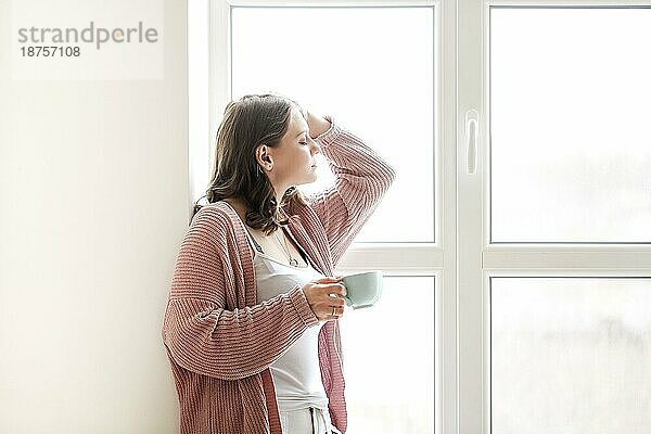 Junge Frau mit Tasse heißem Getränk  die den Kopf berührt und sich in der Nähe des Fensters am sonnigen Morgen zu Hause entspannt