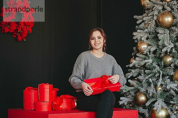 Studiofoto von jungen schönen Frau mit Abendmakeup sitzt neben Xmas Tree mit goldenen Kugeln geschmückt  glücklich Dame mit Neujahrsgeschenke in festlichen leuchtend roten Geschenkpapier umgeben