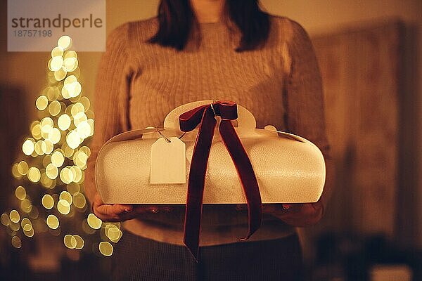 Ausgeschnittenes Foto einer nicht erkennbaren Frau  die eine Weihnachtsgeschenkbox hält  während sie zu Hause mit einem unscharfen Weihnachtsbaum im Hintergrund steht. Selektiver Fokus einer Frau  die ein Neujahrsgeschenk erhält