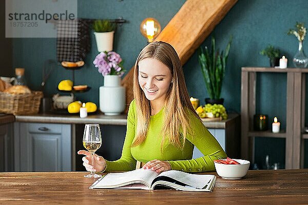 Junge Frau liest eine interessante Zeitschrift an einem Holztisch mit Obst und Wein an einem Wochenende zu Hause