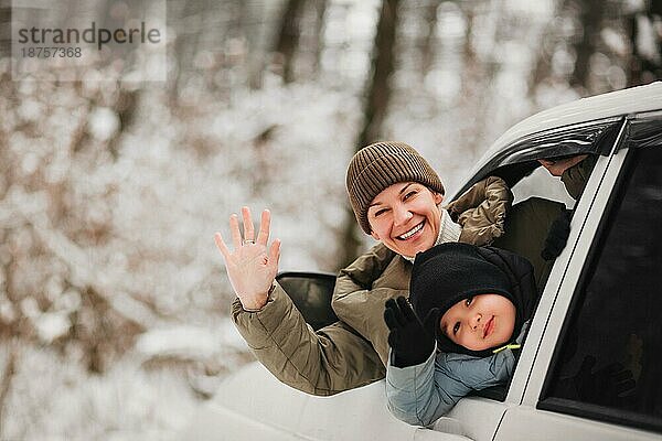 Fröhliche erwachsene Frau und kleiner Junge in Oberbekleidung  die in die Kamera schauen und mit den Händen winken  während sie aus dem Fahrzeugfenster auf einem unscharfen Hintergrund eines Winterwaldes schauen