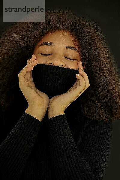 Zufriedene afroamerikanische lockige Frau  die die Augen schließt und das Gesicht mit einem warmen schwarzen Pulloverkragen bedeckt  vordunklem Studiohintergrund  lächelnde Frau  die sich im Rollkragenpullover aufwärmt. Vertikales Foto