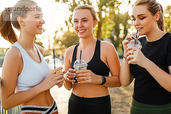 Junge Frau in Sportkleidung lächelt und schaut weg  während sie ein frisches gesundes Getränk in der Nähe von Freundinnen genießt