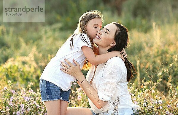 Zarte Tochter küsst entzückte Mutter auf die Wange  während sie sich auf einer blühenden Sommerwiese ausruht