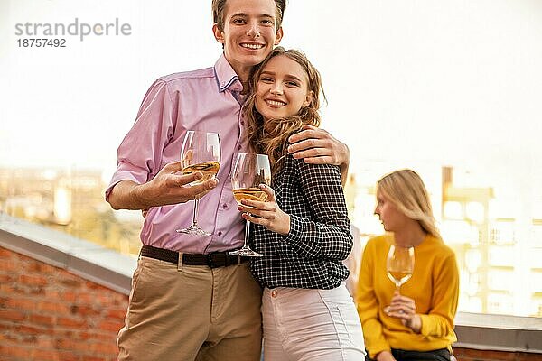 Glückliches junges verliebtes Paar  das sich auf der Terrasse eines Hauses umarmt  Gläser mit Weißwein in der Hand  lächelnd und mit der Kamera posierend  Mann und seine geliebte Frau  Zeit mit Freunden auf einer Sommerparty verbringen