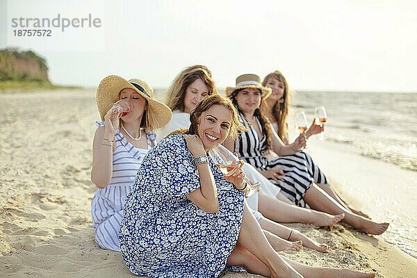 Gruppe von begeisterten Freundinnen  die Wein genießen und miteinander reden  während sie sich am Sandstrand in der Nähe des wogenden Meeres im Sommer entspannen