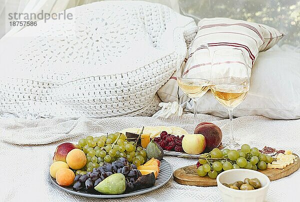 Paar Gläser mit Wein in der Nähe von Tischen mit verschiedenen frischen reifen Früchten auf Decke mit Kissen während romantisches Picknick im Garten platziert