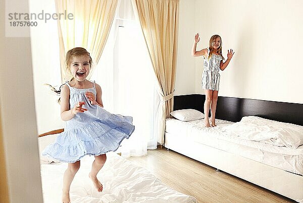 Zwei niedliche sorglos kleine Schwestern Mädchen in lässige Kleidung spielen  die Spaß in Kinderzimmer in sonnigen Morgen. Loving freudige Kinder springen auf Betten zu Hause  Kinder verbringen glückliche Freizeit Indoor