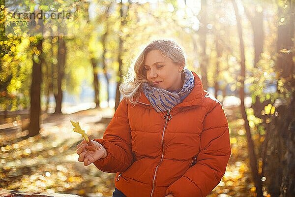 Zufriedene Frau in Oberbekleidung lächelt und betrachtet ein trockenes Blatt an einem sonnigen Wochenendtag im Herbstpark