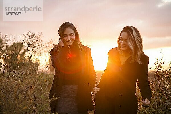 Zwei glückliche Frauen  die sich an den Händen halten  spazieren durch ein herbstliches Feld bei Sonnenuntergang und lächeln  sorglose beste Freundinnen  die einen Abendspaziergang auf einer Wiese auf dem Lande genießen