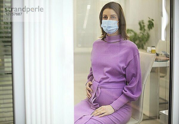 Junge schöne schwangere Frau in medizinischen chirurgischen Maske und sehr perri Kleid in Quarantäne zu Hause am Fenster. Coronavirus  Virus  Isolation  zu Hause bleiben