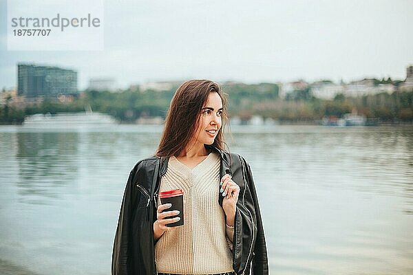 Junge Frau mit Kaffee zum Mitnehmen in der Hand in lässiger Kleidung am Flusswasser am Herbsttag mit Stadtlandschaft im Hintergrund  romantisch träumende Frau an der Küste  die Erholung in der Natur genießt