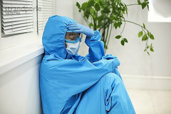 Müde Ärztin in persönlicher Schutzausrüstung (PSA)  deprimiert nach schwieriger  stressiger Schicht während der Covid 19 Pandemie  sitzt auf dem Boden im Krankenhaus. Überfordertes Gesundheitspersonal