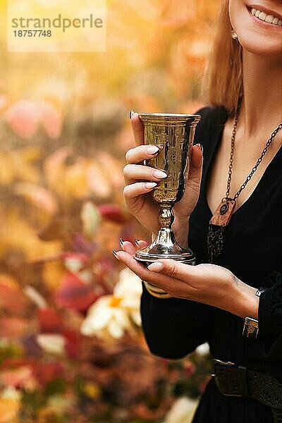 Cropped Schuss der jungen fröhlichen Frau in schwarzem Kleid hält Vintage Metall Glas in den Händen  während im Herbst Park mit bunten Baum Blätter auf Hintergrund  trinken Wein auf Picknick im Wald stehen