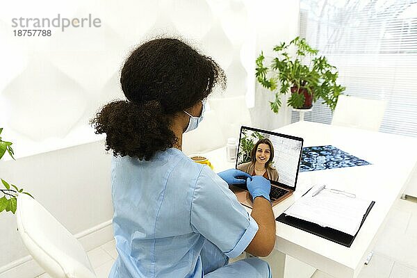 Aufnahme einer attraktiven afroamerikanischen Ärztin  die einem Patienten über einen Videoanruf mit Laptop in der Sprechstunde die medizinische Behandlung erklärt