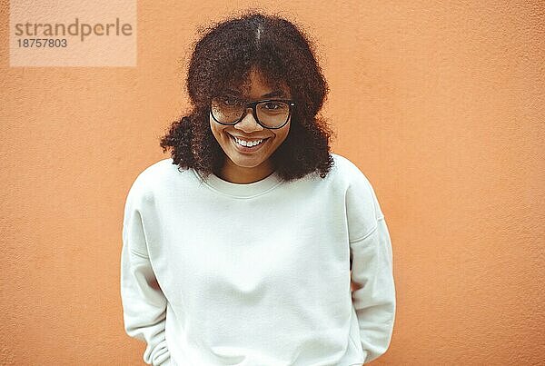 Fröhliches  positives  junges afroamerikanisches Mädchen mit Brille  lächelnde Afroamerikanerin  die eine Brille hält und mit den Händen das Gestell begradigt  breit lächelnd in die Kamera schaut  im Freien an einer orangefarbenen Wand stehend