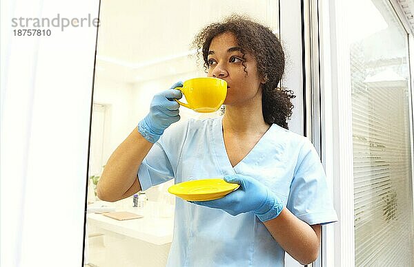 Junge afroamerikanische Ärztin in Uniform und Handschuhen  die Kaffee oder Tee trinkt  während sie eine Pause zwischen den Patientengesprächen in der Klinik einlegt  medizinische Mitarbeiterin  die sich während eines harten Arbeitstages im Krankenhaus ausruht