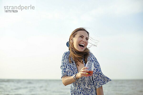 Fröhliche adultes Weibchen in Sommerkleidung lächelt und schaut weg  während sie am Sandstrand in der Nähe des Meeres Wein genießt