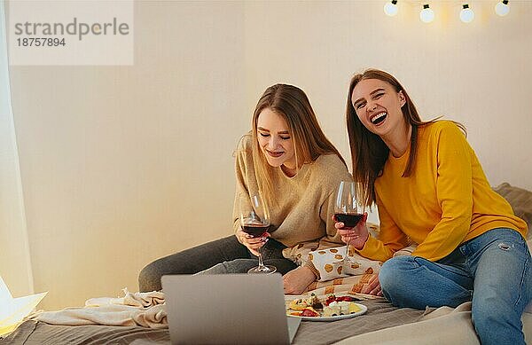 Zwei süße  fröhliche  lustige Freundinnen  die zusammen Spaß haben  einen Heimkinoabend veranstalten  Komödien auf dem Laptop anschauen  laut lachen  auf dem Bett sitzen mit einer Käseplatte und einem Glas Rotwein