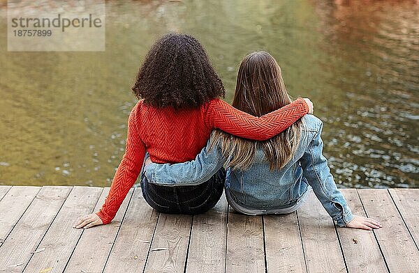 Rückenansicht von zwei glücklichen multirassischen Freundinnen sitzen in der Nähe von Fluss mit Glück  zwei beste weibliche Freunde der verschiedenen Rassen entspannen in der Nähe von Teich im Herbst Wochenende Tag