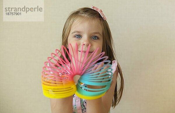 Cute adorable kleines Mädchen spielt mit Kunststoff Regenbogen magische Feder  hält bouncy dehnbar Spielzeug und lächelt in die Kamera. Glückliches Kind vorbeige Hintergrund genießen Freizeit zu Hause