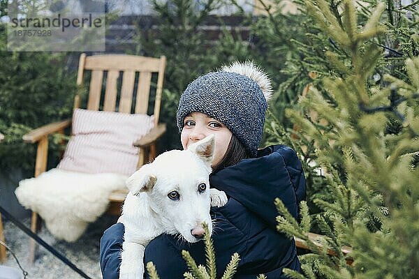 Lächelndes Teenagermädchen in warmer Kleidung  das ein süßes weißes Hündchen in den Armen hält  während es in einem Hinterhof mit Nadelbäumen sitzt  den Hund umarmt und das Winterwetter in Gesellschaft seines besten Freundes genießt