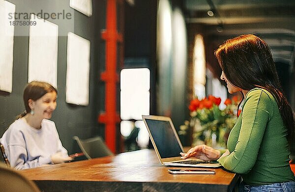Seitenansicht einer glücklichen Frau  die lächelt und auf ihrem Laptop surft  während sie am Tisch sitzt und an einem Fernprojekt in der Cafeteria arbeitet