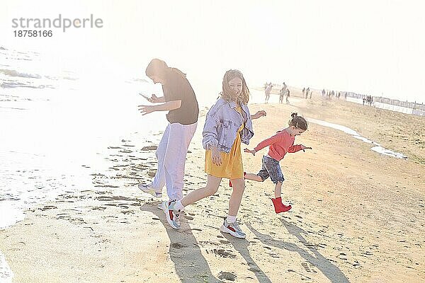 Foto von hinten von einer fröhlichen Familie  die am Strand steht und auf die Brandung und die Wellen schaut  Mutter und zwei Töchter verbringen Zeit am sonnigen Sommerstrand  die kleine Schwester läuft herum und klatscht vor Freude in die Hände