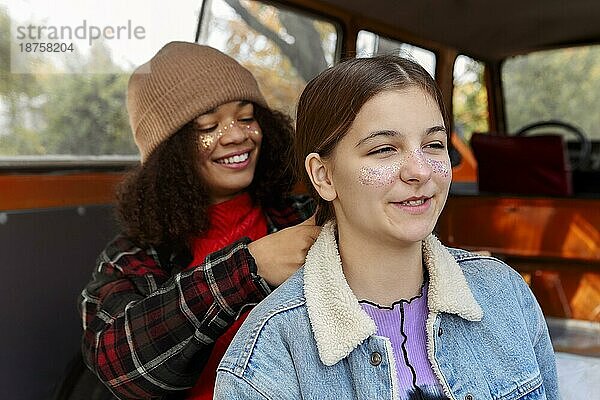 Verschiedene Freundinnen sitzen auf dem Kofferraum eines Autos im Wald  verbringen Zeit miteinander in der Natur  junge lächelnde afroamerikanische Frau flechtet die Haare ihrer besten Freundin  während sie mit einem Minivan unterwegs ist