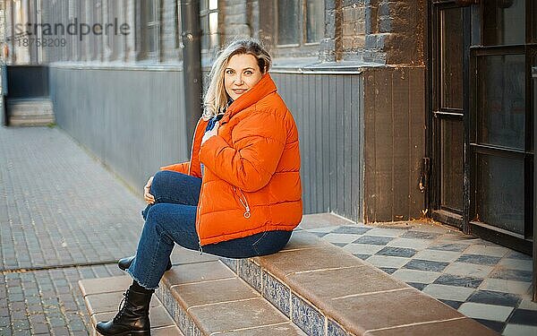 Glücklich lächelnde Frau mittleren Alters in orangefarbener Daunenjacke  die auf einer Betontreppe im Freien sitzt und mit positivem Gesichtsausdruck in die Kamera blickt  während sie sich an einem kalten Herbsttag in der Stadt ausruht