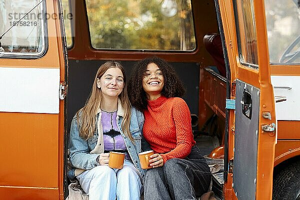 Zwei glückliche multirassische Freundinnen  die im Freien sitzen  heißen Tee trinken und sich bei einem Treffen im Park an einem Herbsttag unterhalten  lächelnde beste Freundinnen  die das Gespräch genießen  während sie draußen sitzen