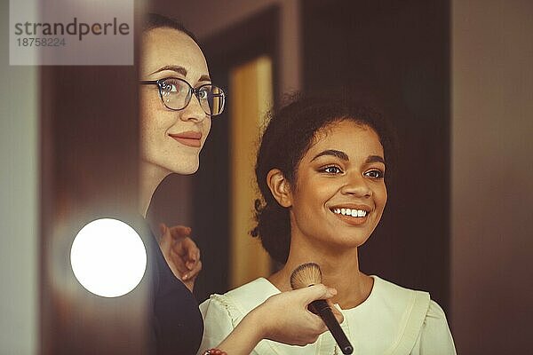 Junge glückliche attraktive afroamerikanische Frau  die einen Visagisten in einem Schönheitssalon besucht  ein professioneller Visagist hält einen Pinsel und trägt Make up auf das Gesicht eines lächelnden schwarzen Mädchens auf  das vor einem Spiegel sitzt