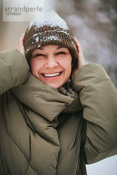 Glücklich lächelnde junge Frau mit Strickmütze und dunkelgrüner gefütterter Jacke  die im Freien in einer verschneiten Winterwaldlandschaft steht  mit positivem Gesichtsausdruck  die das schöne Wetter genießt