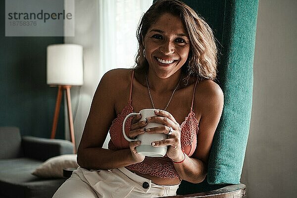 Glückliche junge Latina Frau lächelt in die Kamera  hält eine Tasse und genießt einen neuen Tag. Gesunder Lebensstil Konzept