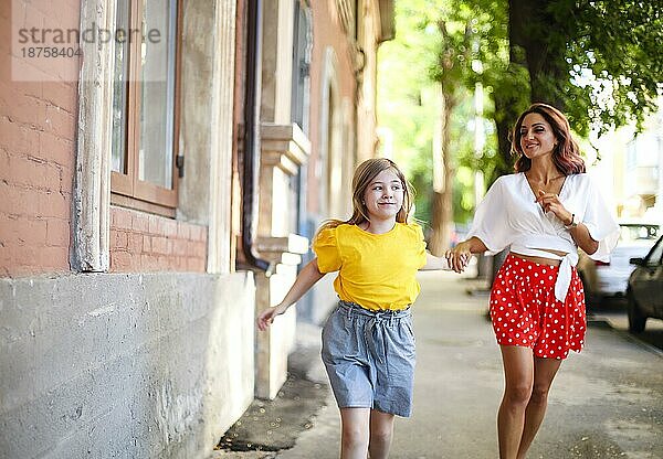 Kleines Mädchen hält die Hand einer stilvollen Frau und geht in der Nähe eines alten Gebäudes an einem sonnigen Sommertag auf einer Straße in der Stadt spazieren