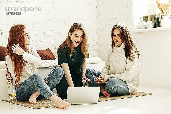 Junge Frauen mit vollem Körperumfang sitzen auf einer Bank und surfen in sozialen Medien auf einem Laptop  während sie sich in einem hellen Raum zu Hause ausruhen