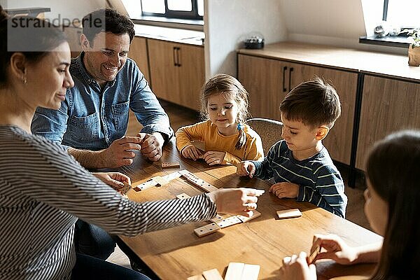Mutter  Vater und Kinder spielen Domino  während sie im Wohnzimmer in der Nähe des Fensters sitzen. Familie verbringt Zeit miteinander