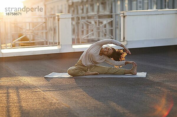 Junge  sportliche  fröhliche  gemischtrassige Frau in Sportkleidung macht Dehnungsübungen  während sie auf einer Yogamatte auf dem Hausdach am frühen Morgen sitzt  fitte Frau  die bei Sonnenaufgang trainiert
