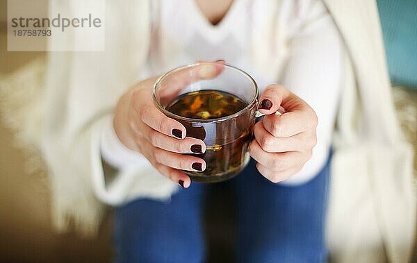 Hohe Winkel der Ernte anonyme weibliche in casual Outfit hält Glas Tasse mit aromatischen Kräutertee beim Entspannen zu Hause