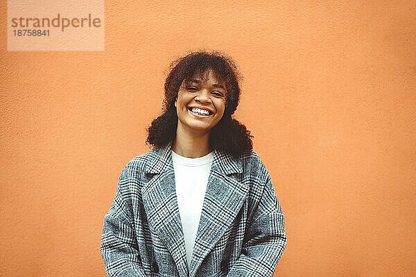 Genießen Sie das Leben. Schöne glückliche afrikanische Ethnizität Mädchen mit üppigen dunklen lockigen Frisur in stilvollen Mantel stehend gegen orange Wand Hintergrund  Blick weg mit breiten hellen Lächeln. Glück Konzept